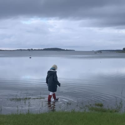 En kvinna med regnrock och gummistövlar vadar vid en havsstrand.