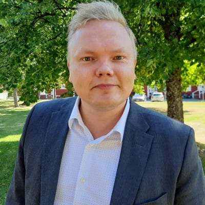 Kulturminister Antti Kurvinen, en blond medelålders man klädd i blå kostym, står under ett träd och tittar in i kameran. 