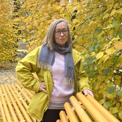Leena Stolzmann har på sig en gul jacka och står omgiven av gula höstlöv. Hon ser allvarlig ut.