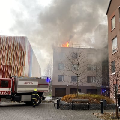 Brandbil framför brinnande höghus. Lågor slår upp genom taket.