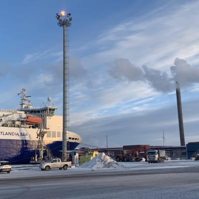 Ett stort containerfartyg står vid hamnkajen med bilar och arbetsmaskiner i förgrunden. 
