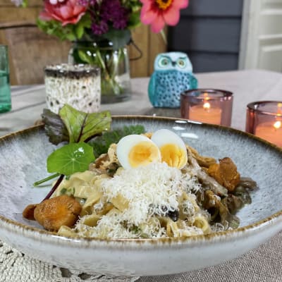 Ett fat med en portion pasta med svampsåsen. Portionen är dekorerad med gröna blad ochovanpå såsen finns det riven ost och två ägghalvor av vaktelägg.