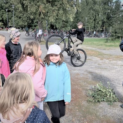En man med bastuhatt spelar gitarr på en skolgård med ettt gäng barn. 