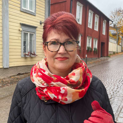 Närbild på en kvinna med rött hår och röd halsduk. Hon tittar in i kameran. Kvinnan bär höstkläder och står på en våt kullerstensgata i Gamla Borgå.