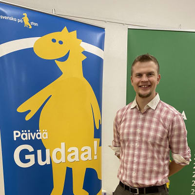 Språkambassadör Jaan Siitonen står bredvid en plansch. Planschen föreställer Svenska veckans maskot, draken Svenni och texten Päivää Gudaa. 