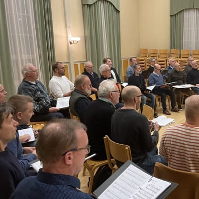En dirigent står framför en flygel och leder en manskör med koristerna sittande på stolar i en sal. 