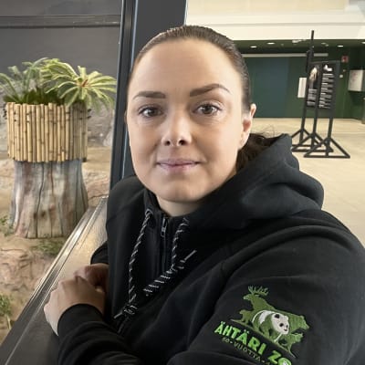 En kvinna med svart munkjacka lutar mot ett staket till en inhängnad med en pandabjörn. 