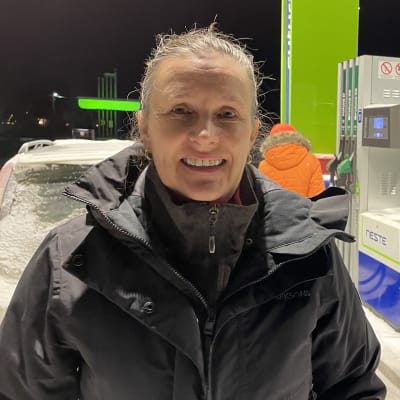 En kvinna med svart jacka står vid en tankstation med en snötäckt bil i bakgrunden. 