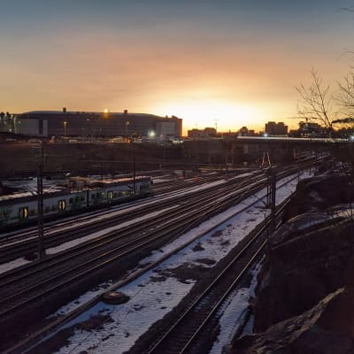 Ett tåg kör i ett vintrigt landskap i Ilmala. I bakgrunden en stigande sol och Helsingfors Arenan.