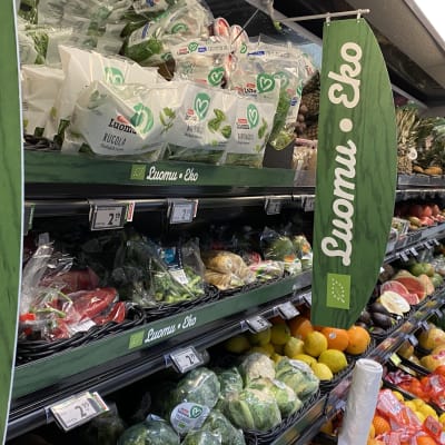 En butikshylla med massor av frukt och grönsaker. På gröna banderoller står det luomu eko.