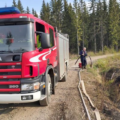 En brandman jobbar med en släckningsslang vid en brandbil. Bilen står parkerad på en skogsväg.