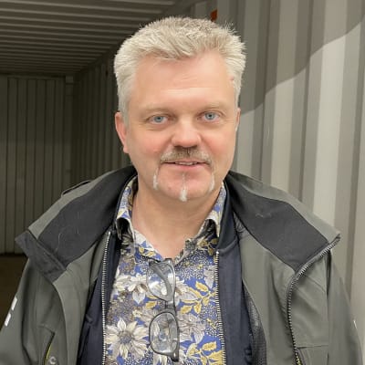 En man med grågrön jacka och blommig skjorta står i öppningen av en stor kontainer. 