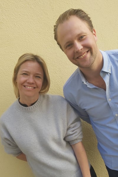 Programledaren Sonja Kailassaari och skådespelaren Dennis Nylund poserar glatt framför en rappad gul husvägg.