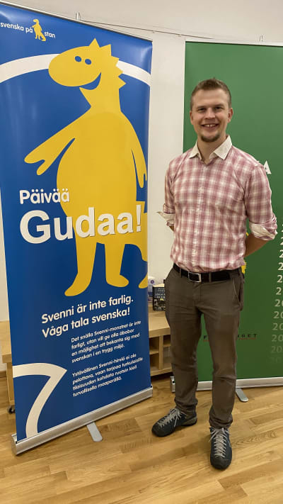 Språkambassadör Jaan Siitonen står bredvid en plansch. Planschen föreställer Svenska veckans maskot, draken Svenni och texten Päivää Gudaa. 