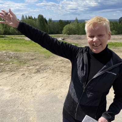 Nainen osoittaa työmaa-alueella oikealla kädellään sivulle ja nauraa kameralle.