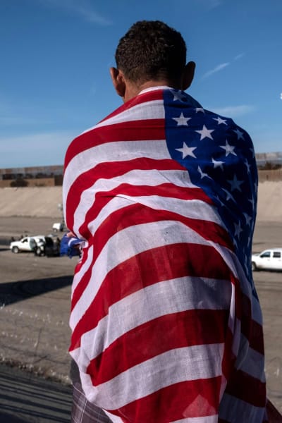 En migrant från Centralamerika insvept i USA:s flagga blickar ut över den torra flodbädden nära gränsövergången El Chaparral, Tijuana vid gränsen mellan USA och Mexiko 