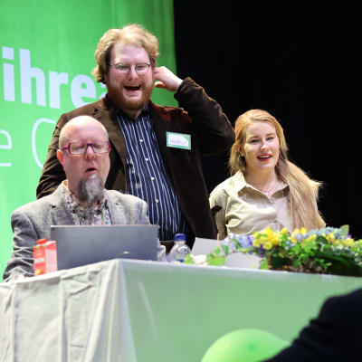 Två män med skägg och en kvinna med långt hår vid ett ordförandebord. Texten Vihreät De Gröna skymtar på en skärm bakom dem..
