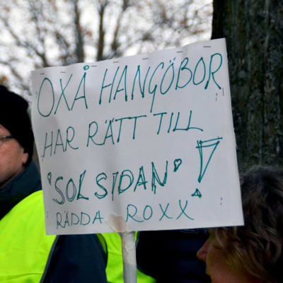 Tre människor i reflexvästar står med i en demonstration. I mitten syns en vit hemgjord skylt där det står Oxå Hangöbor har rätt till "Solsidan", rädda Roxx!