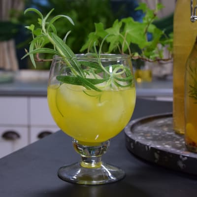 En neongul drink med is och en kvist rosmarin i ett glas på fot.