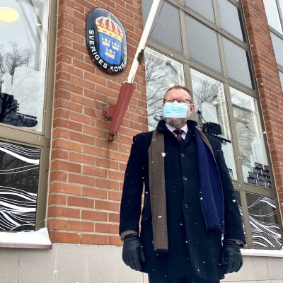 En man i vinterkläder står utanför en byggnad. På väggen finns ett emblem där det står Sveriges konsulat.