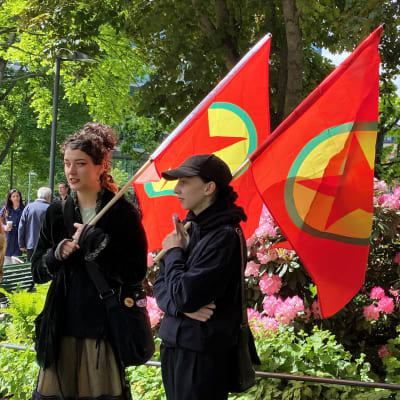 Två kvinnor håller i PKK-flaggor på en demonstration utomhus vid en park.