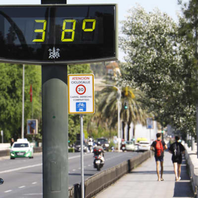 En skylt vid en väg visar att det är 38 grader varmt. Bilar kör på vägen och människor går på trottoaren.