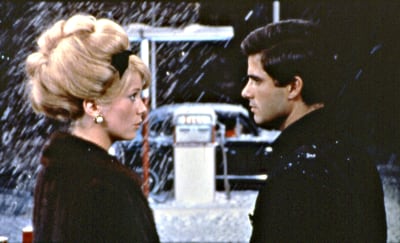Nainen ja mies katsovat toisiaan, taustalla öinen huoltoasema ja lumisadetta, kuva elokuvasta Cherbourgin sateenvarjot.
