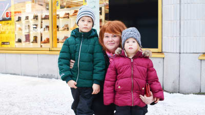 En kvinna sitter på knä och håller armarna om två barn, en flicka och en pojke. Familjen är utomhus i en snöig stadsmiljö.
