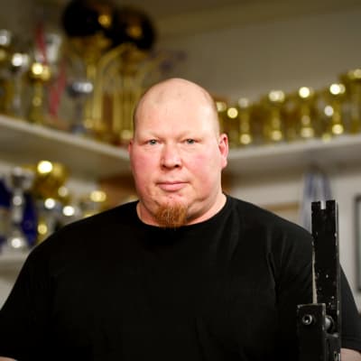 Ove LEhto poserar framför en enorm rad av pokaler och priser i sitt gym
