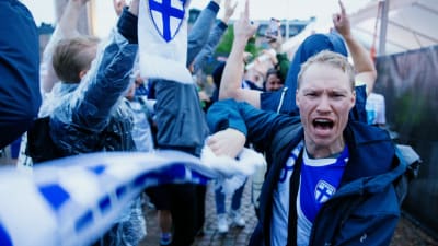 Väldigt glada fans. En man står och viftar en Finland-halsduk och ser ut att skrika något i förgrunden.
