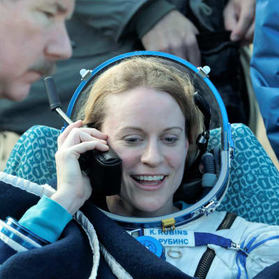 Kate Rubins puhuu satelliittipuhelimeen laskeutumisen jälkeen. Hänellä on yllään avaruuspuku. Ympärillä hyörii ihmisiä.