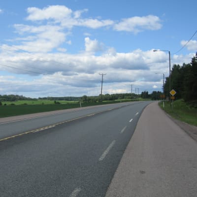 En asfalterad väg med åker på ena sidan och träd på andra. Trafikmärke för förkörsrätt. Telefonstolpar. Sommar, blå himmel med moln.