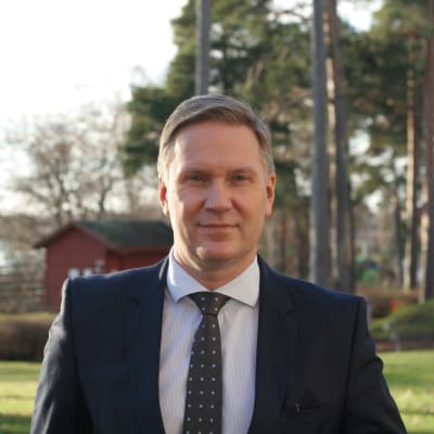 Man i kostym står i en trädgård.
