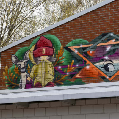 Koulun tiiliseinään on maalattu graffiti, jossa pipopäinen tyttö on kauriin kanssa.