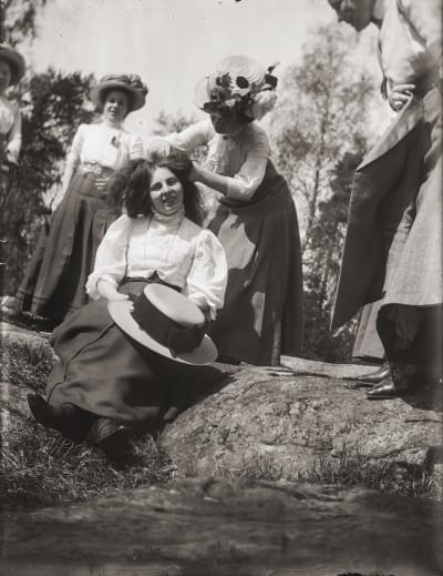 Unga damer från sekelskiftet 1800-1900 lagar varandras hår ute i naturen. 