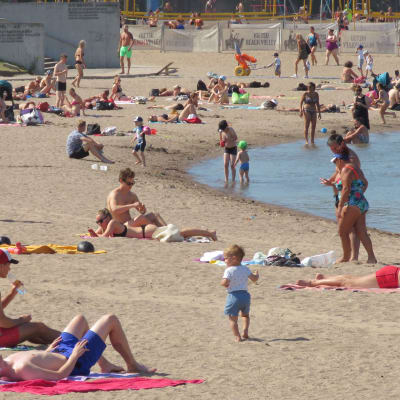 Paljon uima-asuisia ihmisiä hiekkarannalla ja rantavedessä. 