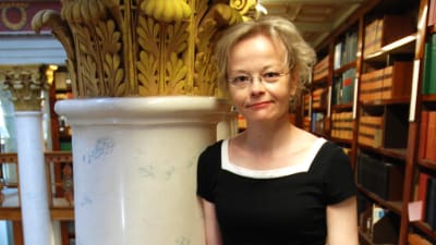 En kvinna iklädd en svart klänning. Står och lutar sig mot ett räcke. Det ser ut som att hon befinner sig i ett bibliotek. 