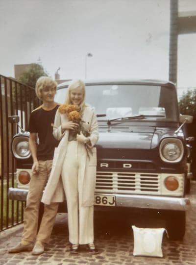 Nuori kihlapari mustan Ford-pakettiauton edustalla, naisella keltainen kukkakimppu sylissä.
