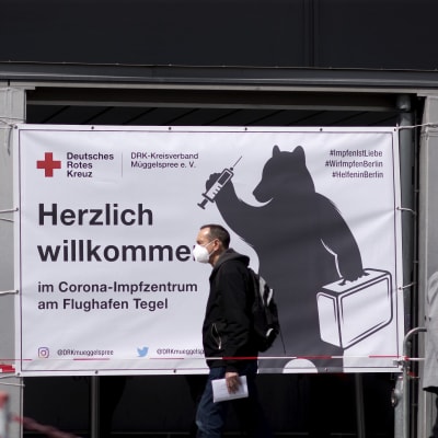 Två män i munskydd går på gatan i Berlin. I bakgrunden syns en reklamskylt för coronavaccin.