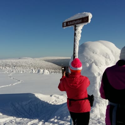 Två skidåkare i rött och lila står på toppen av ett fäll i Lappland och tar foton.