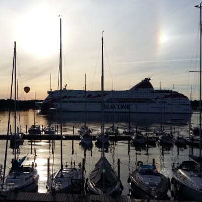 Passagerarfartyget M/S Baltic Princess passerar Airisto Segelsällskaps klubbhamn på Backholmen i Åbo, med segelbåtar i hamnen och en luftballong i bakgrunden.