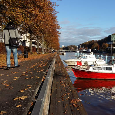 En man går förbi en knallröd båt i Aura å en solig höstdag i Åbo.