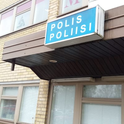 En blå skylt med texten Polis Poliisi ovanför en dörr på ett hus med gula tegel.
