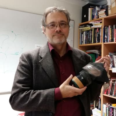 Professor Mats FRidlund, med gasmask  handen, framför sin bokhylla
