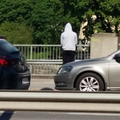 Ung man i ljus munkjacka står på Domkyrkobron och blickar ut över Aura å. Två bilar susar förbi honom