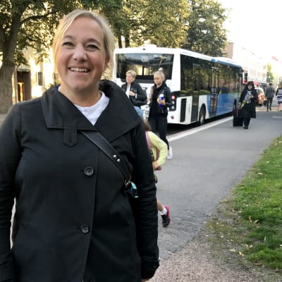 Silja Lindblad står vid busshållplats, i bakgrunden människor och buss.