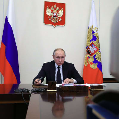 Vladimir Putin sitter vid ett mörkt och blankt skrivbord och tittar in i en videokamera.