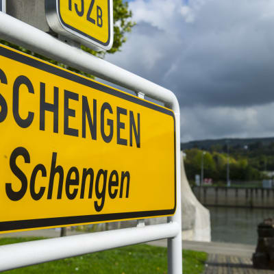 Vasemmalla etualalla isona keltainen Luxemburgissa kuvattu Scengen-kyltti, jonka oikealla puolella sumeaa maisemaa.