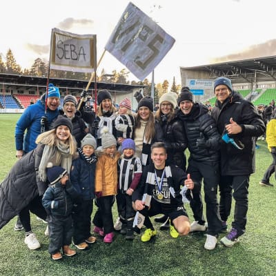 En stor grupp människor i olika åldrar står samlade på en fotbollsplan och omringar fotbollsspelaren Sebastian Strandvall. De småler mot kameran och håller i skyltar där det står "Seba" och "VPS".