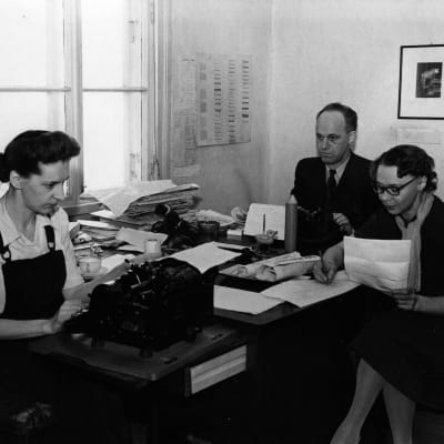 Tre personer, två kvinnor och en man, runt ett bord på en redaktion. Svartvit bild.